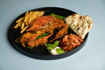 Indian-style Tawa Crab Masala served on a banana leaf in a black plate, accompanied by fresh salad, tangy dip, crispy fries, and soft kulchas on a clean blue background.
