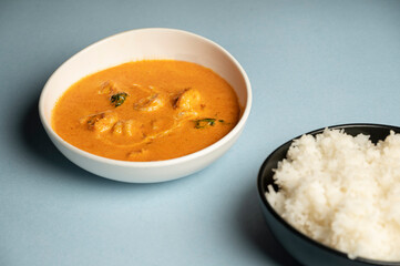 Prawns Thai red curry served in a white bowl and jasmine rice in a black bowl on a blue background. A rich, aromatic curry made with Thai spices and coconut milk.