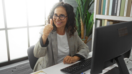 Woman smiling and giving thumbs-up gesture in an office setting, surrounded by books and computer,...