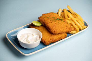 Crispy fish fillet battered and crumb-fried, served with golden fries and tartare sauce on a blue plate placed over a blue background.