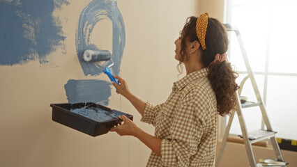 Woman painting wall with blue paint in new home interior with roller and tray, showcasing home improvement in bright, natural light setting.