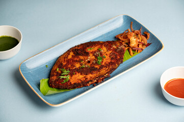 Indian tawa masala kingfish served with fresh salad in a blue plate on banana leaf, accompanied by green and red chutneys in white bowls, styled on a bold blue background.