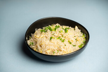 Aromatic egg fried rice served in a black bowl, placed on a solid blue background, showcasing golden egg bits, fresh greens, and a savory, pan-tossed texture.