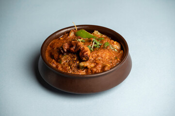 Traditional Goan chicken xacuti served in a rustic brown clay pot, rich with roasted coconut and spices, placed on a solid blue background for a vibrant contrast.