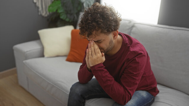 Young man sits contemplatively in a modern living room with neutral decor, conveying an introspective mood in a cozy indoor setting.