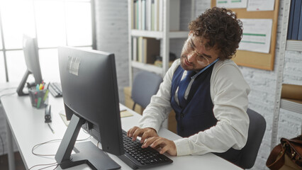 Young hispanic man in office multitasking, typing on computer while talking on phone, capturing a busy work environment with modern interior and professional ambiance.
