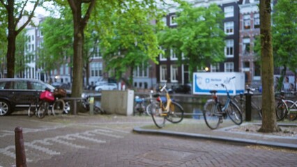 Blurred bicycles and cars along a canal in amsterdam with lush green trees and historic buildings creating a vibrant urban scenery.