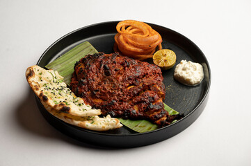 Tandoori pomfret served with salad, dip, sauce, and kulchas on a black plate over white background, styled with vibrant Indian elements and clean presentation.	