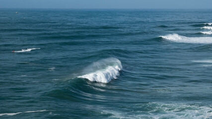 Powerful ocean waves crashing and creating white foam on a sunny day with some boats sailing in the distance, creating a beautiful seascape