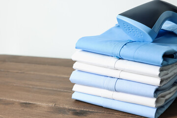 Upright steamer and stack of folded shirts on wooden table near white wall indoors, closeup. Space...