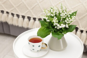 Bouquet of beautiful lily-of-the-valley in vase and cup of tea on coffee table indoors