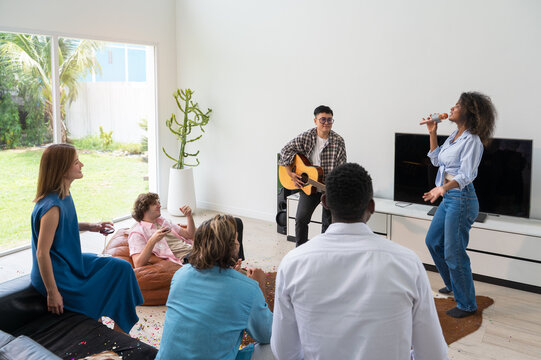 Multiethnic friends enjoy home karaoke in bright living room. Woman with curly hair sing while female in plaid play guitar while others watch drink red wine. Weekend party at home concept