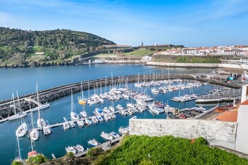 Fototapeta premium Scenic aerial view of marina with sailboats and yachts in natural harbor. Angra do Heroismo marina, historic fortress on Monte Brasil green hillside under blue sky, Terceira Island, Azores, Portugal.