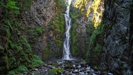 Tall waterfall cascading through narrow rocky gorge covered with moss and greenery, tranquil atmosphere and gentle water flow in lush natural setting