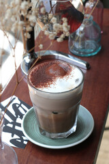A close-up shot of a hot chocolate drink, topped with frothy milk and a dusting of cocoa powder, served in a glass on a green saucer with decorative elements in the background
