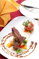 Exquisitely plated gourmet meat dish, possibly beef or lamb, accompanied by colorful sautéed vegetables, a savory sauce drizzle, and garnished with a roasted tomato and mushrooms, presented on a white