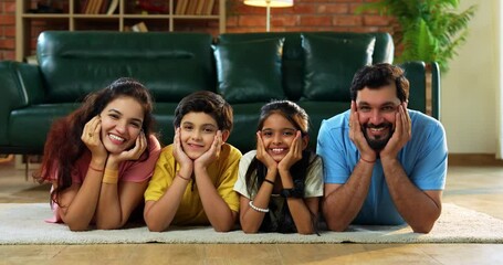Family lying hands on chin posing happily on carpet as Indian father mother son daughter look at camera in modern lavish living room indoors, relaxing together at home for bonding and fun moments
