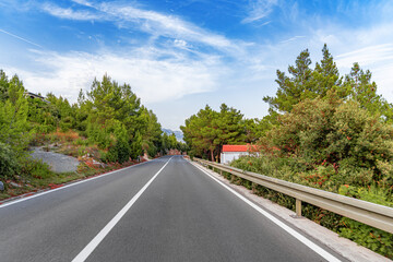A peaceful countryside road stretches ahead through lush greenery and pine trees under a clear blue sky with scattered clouds.