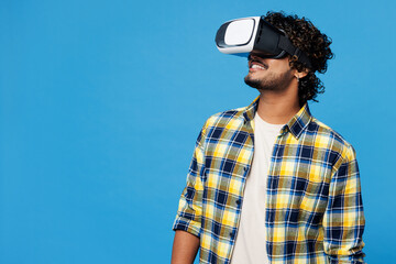 Young smiling fun happy cheerful Indian man he wearing yellow checkered shirt casual clothes watching in vr headset pc gadget isolated on plain blue cyan background studio portrait. Lifestyle concept.