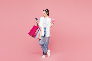 Fototapeta premium Full body young smiling happy woman wear white clothes hold shopping paper package bags use mobile cell phone drink coffee isolated on plain pink color background. Black Friday sale buy day concept.