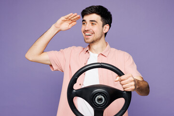 Young smiling happy man he wearing pink shirt t-shirt casual clothes hold steering wheel driving car look far away isolated on plain pastel light purple background studio portrait. Lifestyle concept.