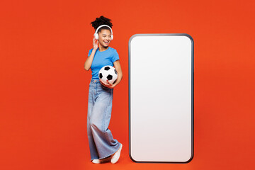 Full body young woman fan wear blue clothes cheer up football sport team hold soccer ball watch tv live stream big blank screen mobile cell phone listen music isolated on plain red orange background.