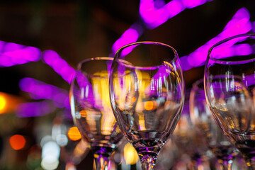 Empty clear wineglasses stand in rows on bar counter at nightclub, blurred purple neon and yellow lights on background. Clean cocktail glasses ready for celebrate party.