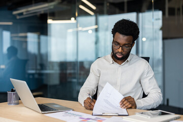 An african american man with glasses working at his desk in a modern office, reviewing documents. A...