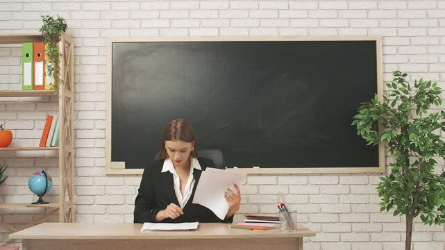 Young woman college teacher at desk in classroom in front of chalkboard reading grading homework or tests of students. Education concept.