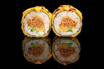 Close-up of a deep fried sushi roll cut in half, filled with cooked salmon, creamy cheese, cucumber and drizzled with sauce. Presented on black background with reflection