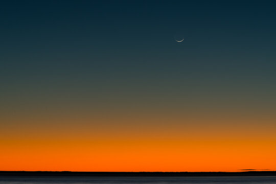 Bright orange and dark blue post sunset sky with crescent moon and low horizon