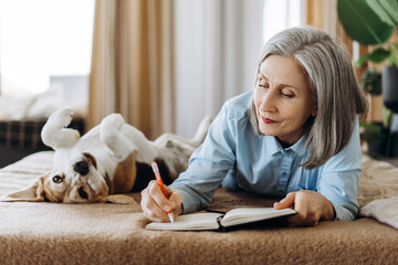 Senior woman writing in notebook with dog on bed
