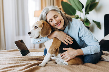 Smiling senior woman hugging beagle dog at home on bed with laptop