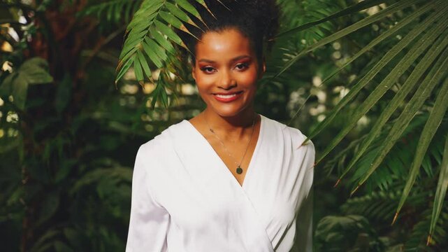 young adult happy African American woman fashion model walking in tropical forest green palm tree, smiling face girl eyes looking at camera with love enjoying summer garden nature tropics. White dress