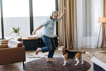 Senior woman listening music with headphones and dancing with beagle dog at home