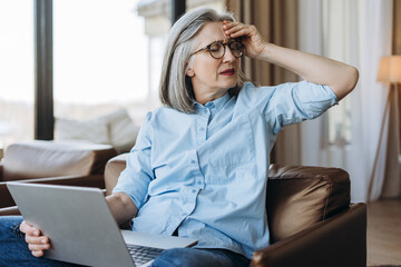 Stressed senior woman suffering headache while working on laptop at home