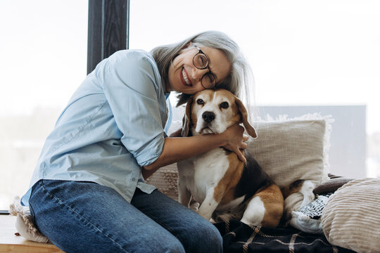 Senior woman hugging beagle dog at home by window - Powered by Adobe
