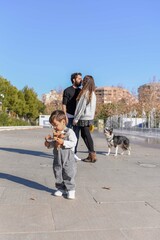Fototapeta premium Vertical image of Toddler boy playing with dry leaves while parents kissing in background at park