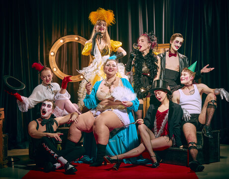 Group of artistic people, cabaret performers in eccentric outfits and makeup, pose on red carpet with dramatic expressions and stage props. Concept of show, circus, retro, vintage, performance.