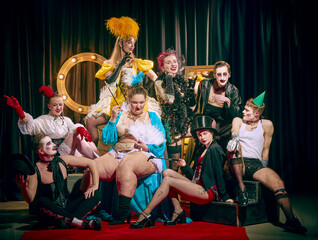 Large troupe of cabaret performers in elaborate vintage costumes pose together on red carpet,...