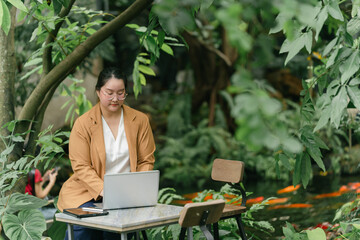Woman working on laptop in lush outdoor setting surrounded by greenery and koi pond, creating...