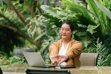 Woman sitting at outdoor workspace surrounded by lush greenery stretches her arms while working on...