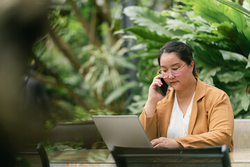 Woman in brown blazer is working on laptop while talking on phone, seated in outdoor garden setting surrounded by lush green plants, creating serene and professional atmosphere