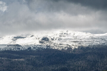 Obraz premium Le sommet d’une montagne enneigée dans le Jura, enveloppé d’une lumière dramatique sous un ciel chargé, offrant un paysage saisissant et contrasté.