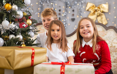 Happy brother with two sisters are sitting on sofa together with gift boxes next to the new year tree