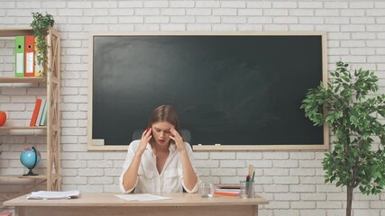 Young woman college teacher at desk in classroom in front of chalkboard grading test papers, stressed expression, having headache and drinks water.