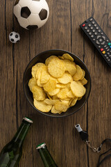 A vertical bowl of potato chips with soccer balls, beer bottles, and a remote set on wooden surface signifying game night essentials