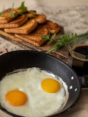Fried eggs in a rustic iron pan, toast on a wooden board and a cup of coffee for breakfast.