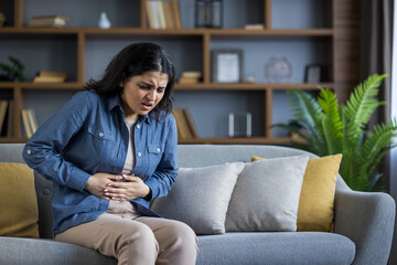 A woman experiencing stomach pain sits on a sofa, expressing her discomfort and holding her stomach.