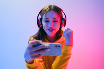 Joyful young woman plays mobile games wearing headphones, celebrating a win under colorful neon...